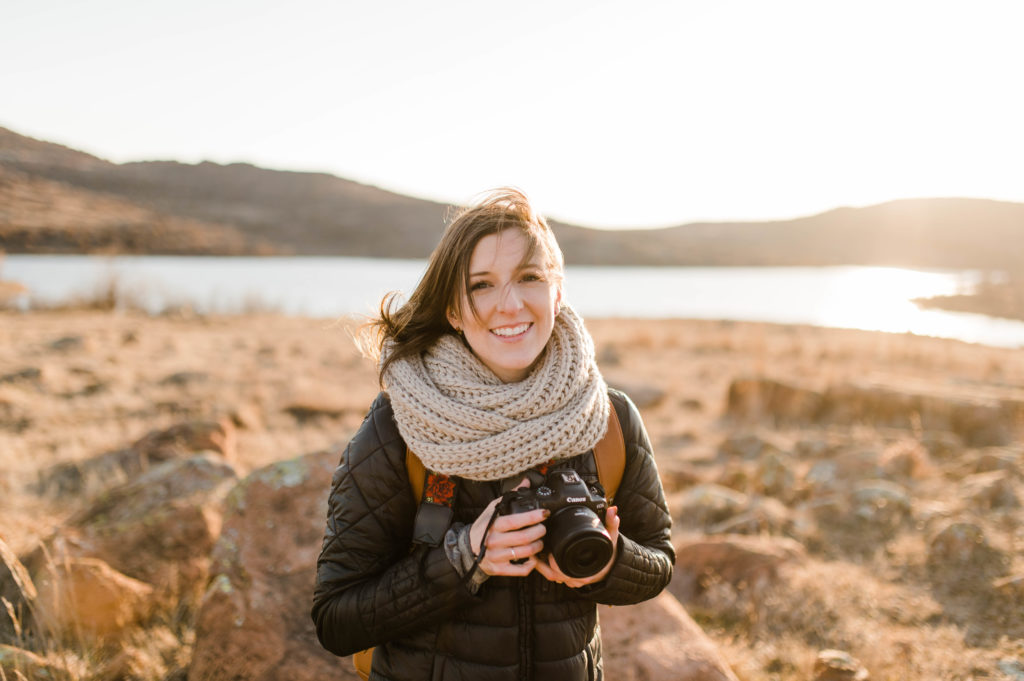 a photographer standing and holding her camera dressed in a puffy jacket and scarf in the background you can see a low mountain skyline with the sun beaming down